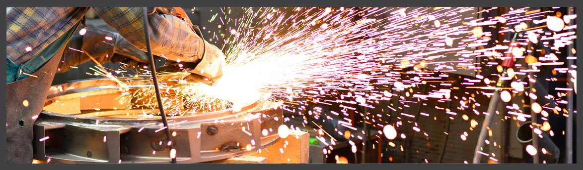 Worker using a grinding tool on a metal component in a foundry, with bright sparks flying outward during the metalworking process.