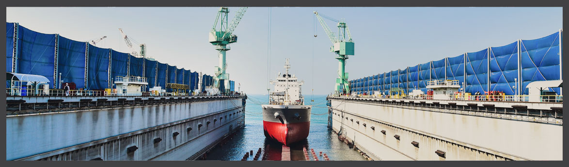Cargo ship docked in a shipyard dry dock with large cranes and blue barriers on either side, under a clear blue sky.
